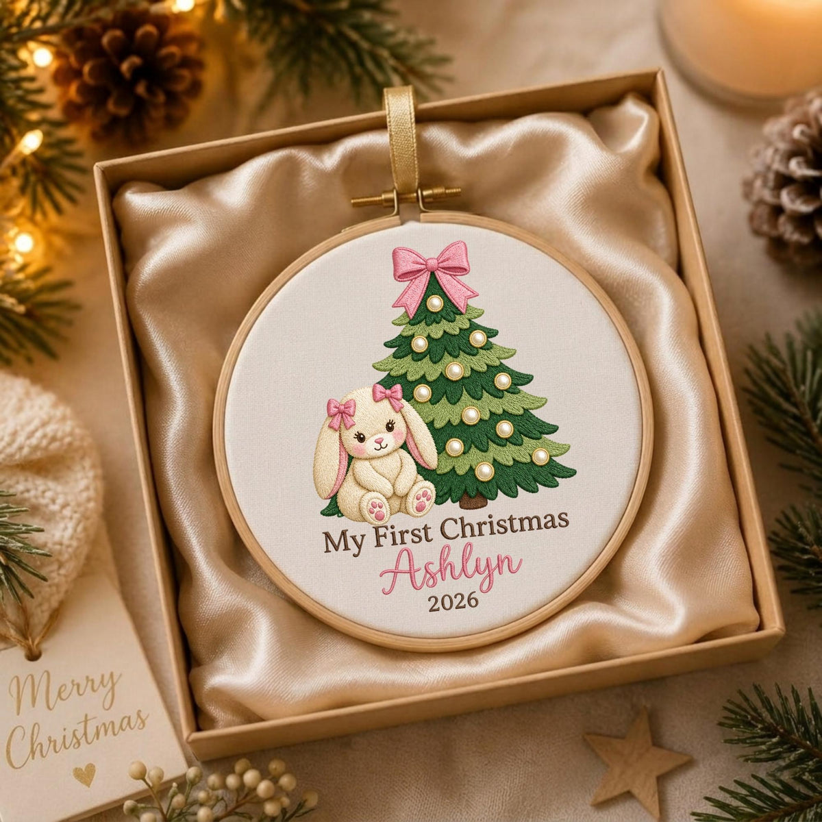 May include: A round wooden embroidery hoop ornament featuring a white fabric center with an embroidered green Christmas tree, a cream-colored bunny with pink bows, and the text My First Christmas Ashlyn 2026, presented in a satin-lined gift box.