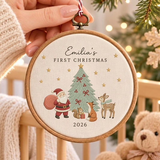 May include: A round wooden embroidery hoop ornament featuring cream fabric with embroidered text reading Emilia's FIRST CHRISTMAS and 2026. The design includes a green Christmas tree, Santa Claus, a squirrel, a fox, and a deer with gold stars.