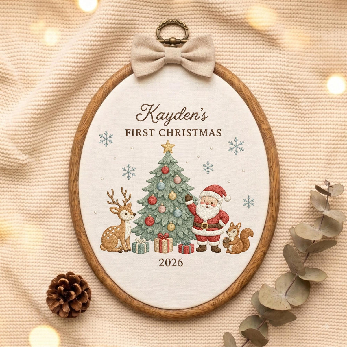 May include: An oval wooden embroidery hoop containing white fabric with the embroidered text Kayden's FIRST CHRISTMAS 2026. The design features a decorated Christmas tree, a deer, Santa Claus, a squirrel, and snowflakes, topped with a beige bow.