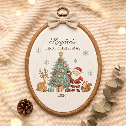 May include: An oval wooden embroidery hoop containing white fabric with the embroidered text Kayden's FIRST CHRISTMAS 2026. The design features a decorated Christmas tree, a deer, Santa Claus, a squirrel, and snowflakes, topped with a beige bow.