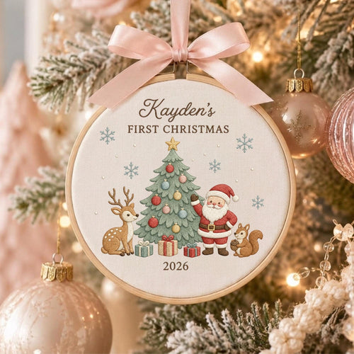 May include: A round wooden embroidery hoop ornament featuring a cream fabric center with a printed design of a Christmas tree, Santa Claus, a deer, and a squirrel. The text reads Kayden's FIRST CHRISTMAS 2026. A light pink satin ribbon bow is attached.