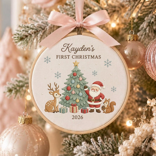 May include: A round wooden embroidery hoop ornament featuring a cream fabric center with a printed design of a Christmas tree, Santa Claus, a deer, and a squirrel. The text reads Kayden's FIRST CHRISTMAS 2026. A light pink satin ribbon bow is attached.