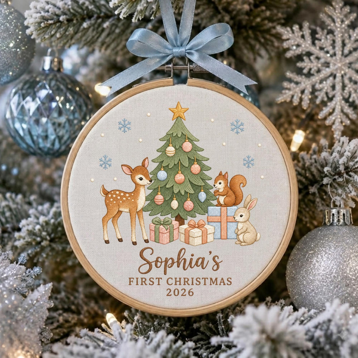 May include: A round wooden embroidery hoop ornament featuring a light fabric background with a printed Christmas tree, a deer, a squirrel, and a rabbit. The text reads Sophia's FIRST CHRISTMAS 2026. A light blue ribbon is tied at the top.