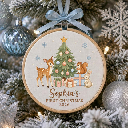 May include: A round wooden embroidery hoop ornament featuring a light fabric background with a printed Christmas tree, a deer, a squirrel, and a rabbit. The text reads Sophia's FIRST CHRISTMAS 2026. A light blue ribbon is tied at the top.