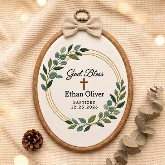 May include: An oval wooden embroidery hoop containing white fabric with a green leaf wreath, a brown cross, and black text reading God Bless, Ethan Oliver, BAPTIZED, and 12.25.2026. A beige fabric bow is attached to the top metal hanging loop.