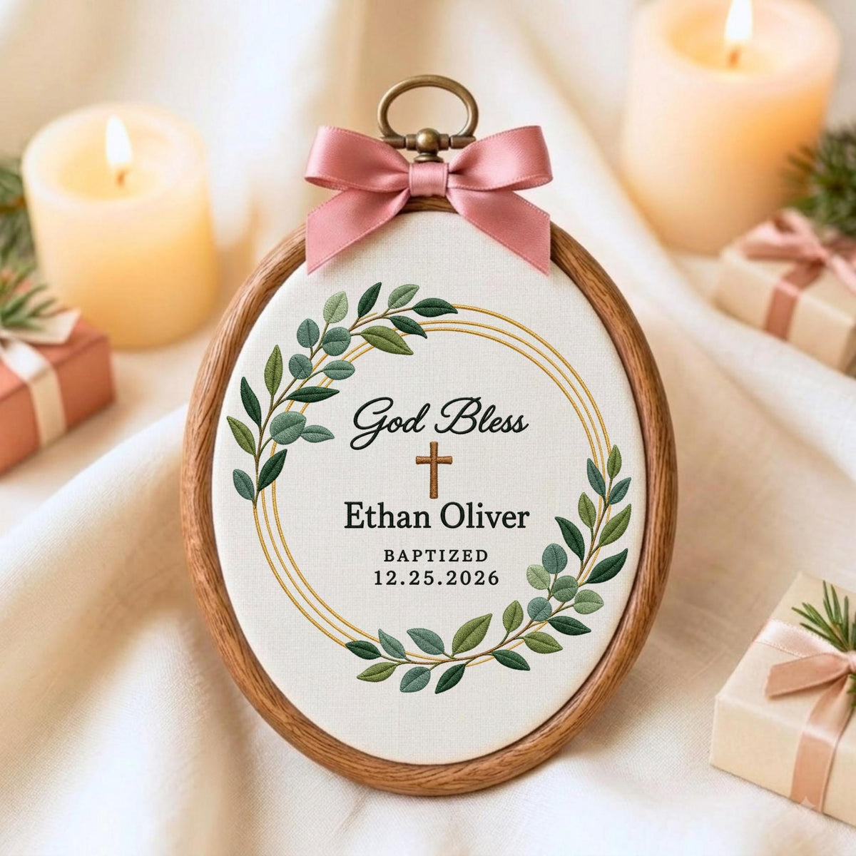 May include: An oval wooden embroidery hoop containing white fabric with a green leaf wreath, a brown cross, and the text God Bless, Ethan Oliver, and BAPTIZED 12.25.2026. A dusty pink satin ribbon bow is attached to the top metal hanging loop.