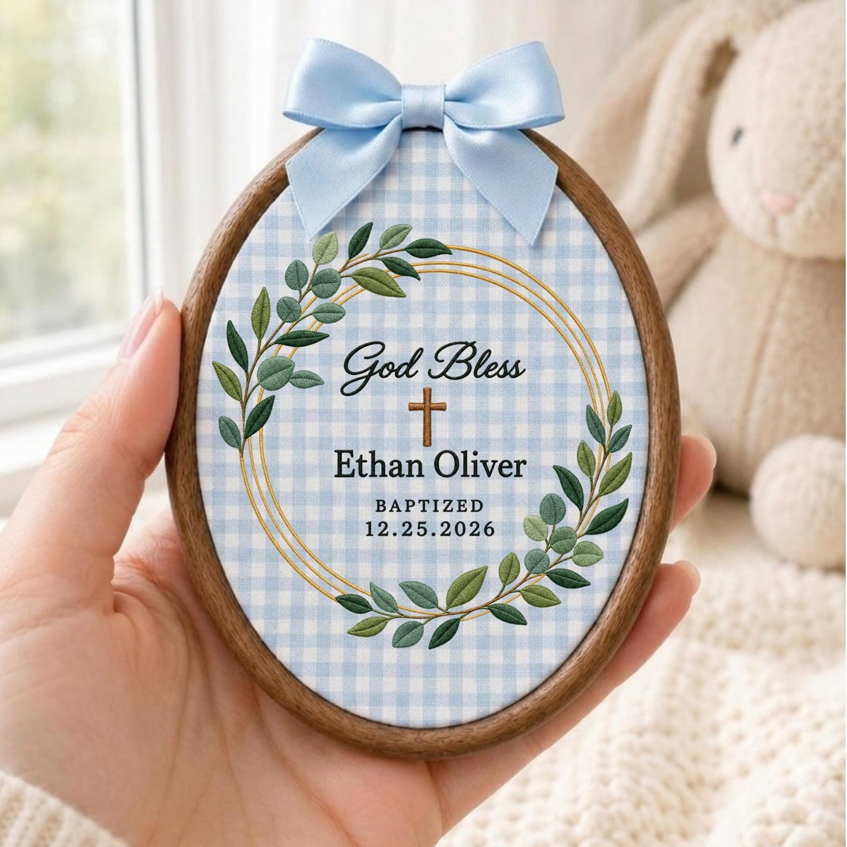May include: An oval wooden embroidery hoop frame containing fabric with a light blue and white gingham pattern. The fabric features an embroidered green leaf wreath, a gold circular line, a brown cross, and the text God Bless Ethan Oliver.