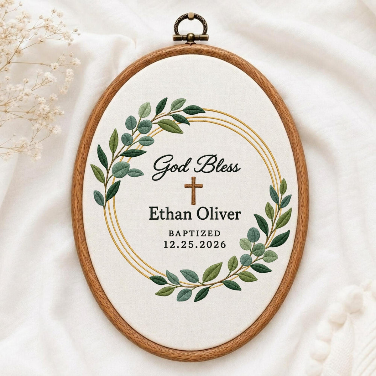 May include: An oval wooden embroidery hoop containing white fabric with a green embroidered leaf wreath, a brown cross, and black text that reads God Bless, Ethan Oliver, BAPTIZED, and 12.25.2026, centered within gold circular lines.