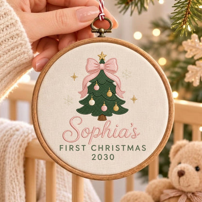 May include: A round wooden embroidery hoop containing off-white fabric with an embroidered green Christmas tree, a large pink bow, and gold and pink ornaments. The text reads Sophia's FIRST CHRISTMAS 2030 in pink and dark green lettering.