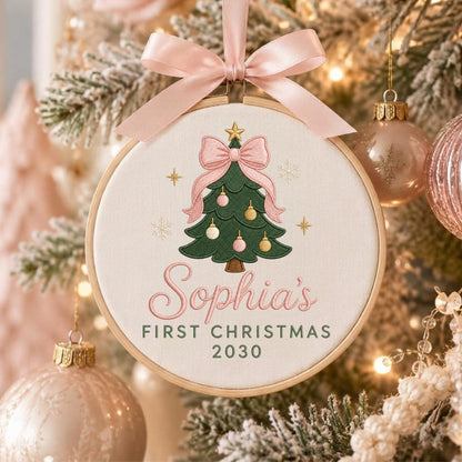 May include: A round wooden embroidery hoop ornament featuring a cream fabric center with an embroidered green Christmas tree, pink bow, and ornaments. The text reads Sophia's FIRST CHRISTMAS 2030. A light pink satin ribbon is tied at the top.