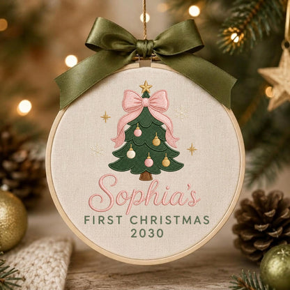 May include: A round embroidery hoop ornament featuring a green Christmas tree with pink and gold ornaments, topped with a pink bow. The text reads Sophia's FIRST CHRISTMAS 2030 in pink and green. An olive green satin ribbon is tied at the top.