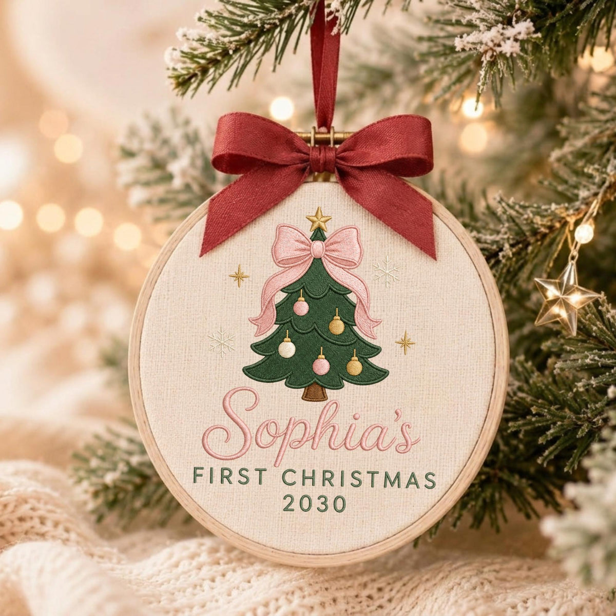 May include: A round wooden embroidery hoop ornament featuring a cream-colored fabric background. The design includes a green Christmas tree with pink and gold ornaments, a large pink bow, and the text Sophia's FIRST CHRISTMAS 2030. A dark red ribbon bow is attached to the top.