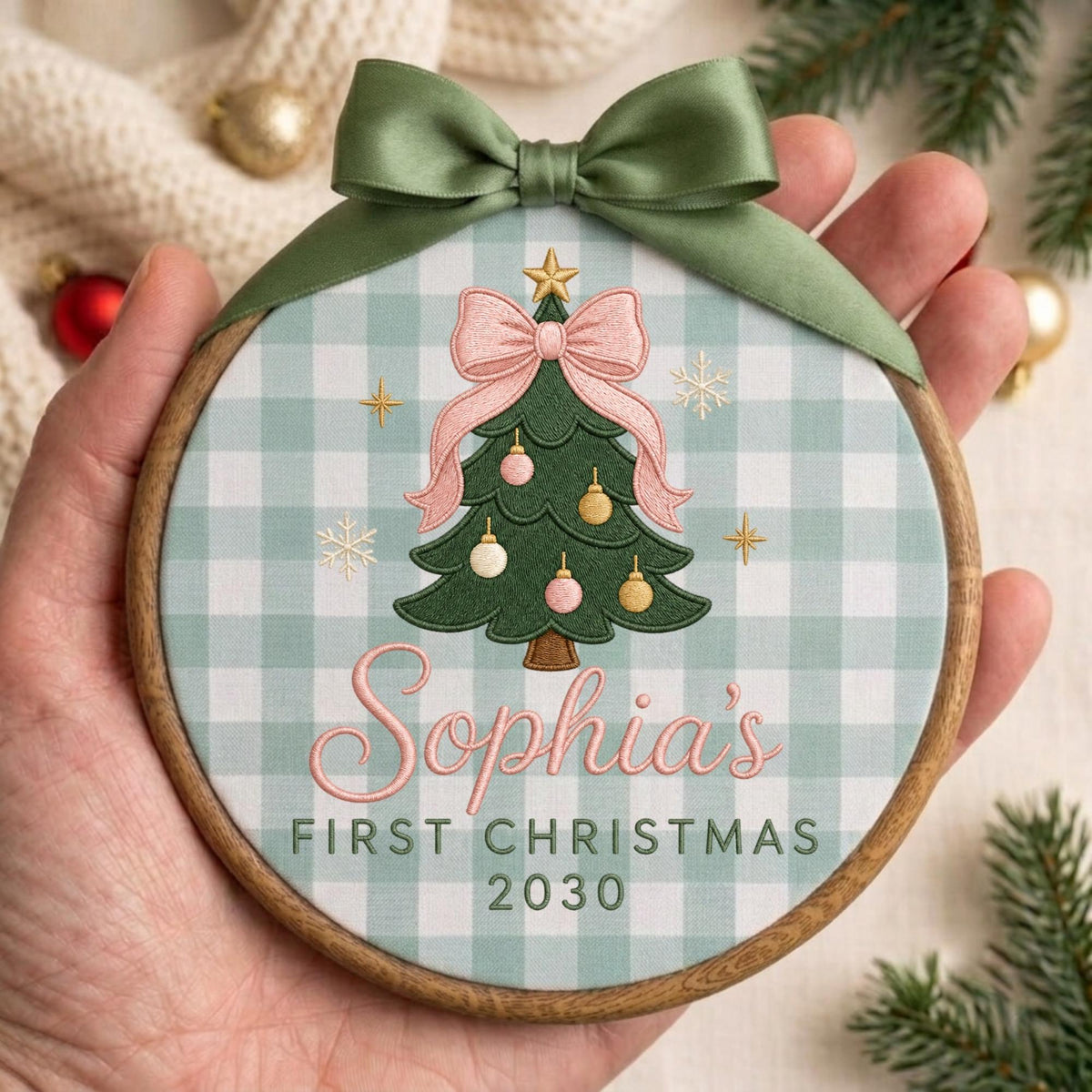 May include: A round wooden embroidery hoop ornament featuring a light green and white gingham fabric background. The center displays a green Christmas tree with pink and gold ornaments, topped with a pink bow and a gold star. Text reads Sophia's FIRST CHRISTMAS 2030. A sage green satin ribbon is tied at the top.