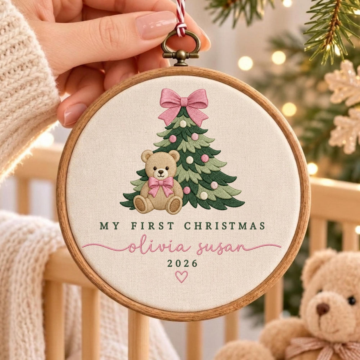 May include: A round wooden embroidery hoop ornament featuring a stitched design of a green Christmas tree with pink ornaments and a pink bow, a light brown teddy bear with a pink bow, and the text My First Christmas, Olivia Susan, 2026.