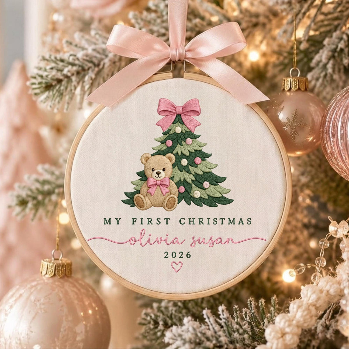 May include: A round wooden embroidery hoop ornament featuring a cream fabric background with a stitched green Christmas tree, a brown teddy bear with a pink bow, and the text My First Christmas, Olivia Susan, 2026, topped with a pink satin ribbon.