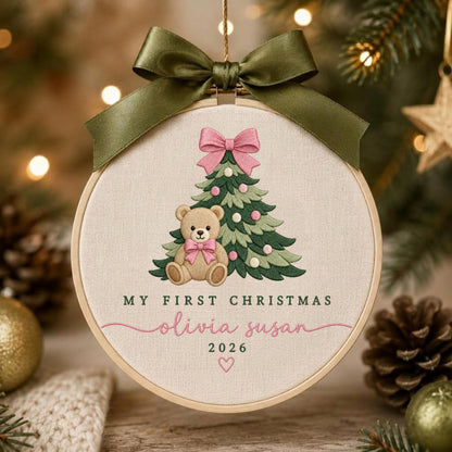 May include: A round embroidery hoop ornament featuring a beige fabric background with a stitched green Christmas tree, a teddy bear with a pink bow, and the text "MY FIRST CHRISTMAS", "olivia susan", "2026", and a small heart, topped with a dark green ribbon.