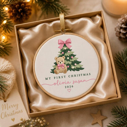 May include: A round wooden embroidery hoop ornament featuring a light brown teddy bear sitting in front of a green Christmas tree with pink ornaments and a pink bow. The text reads My First Christmas, Olivia Susan, 2026, with a small pink heart.