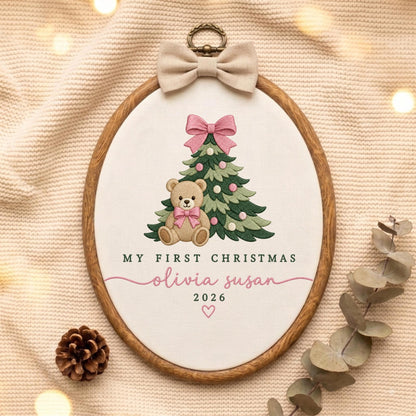 May include: An oval embroidery hoop featuring a tan teddy bear with a pink bow sitting in front of a green Christmas tree with pink ornaments. The text reads MY FIRST CHRISTMAS, olivia susan, and 2026 with a small pink heart below.