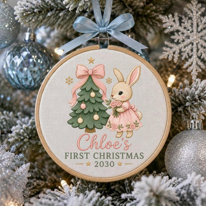 May include: A round wooden embroidery hoop ornament featuring a light-colored fabric background. The design shows a green Christmas tree with pink ornaments and a pink bow, a rabbit in a pink dress holding flowers, and the text Chloe's FIRST CHRISTMAS 2030. A light blue ribbon is tied at the top.