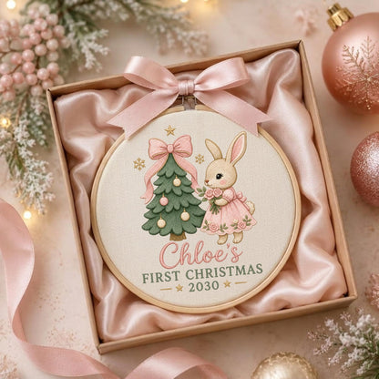 May include: A round wooden embroidery hoop featuring a printed illustration of a rabbit in a pink dress holding flowers next to a decorated Christmas tree. The text reads Chloe's FIRST CHRISTMAS 2030. A pink satin ribbon is tied at the top.
