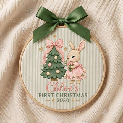 May include: A circular embroidery hoop ornament featuring a green striped fabric background, a dark green satin bow at the top, a green Christmas tree with pink ornaments, and a rabbit in a pink dress. Text reads: Chloe's FIRST CHRISTMAS 2030.