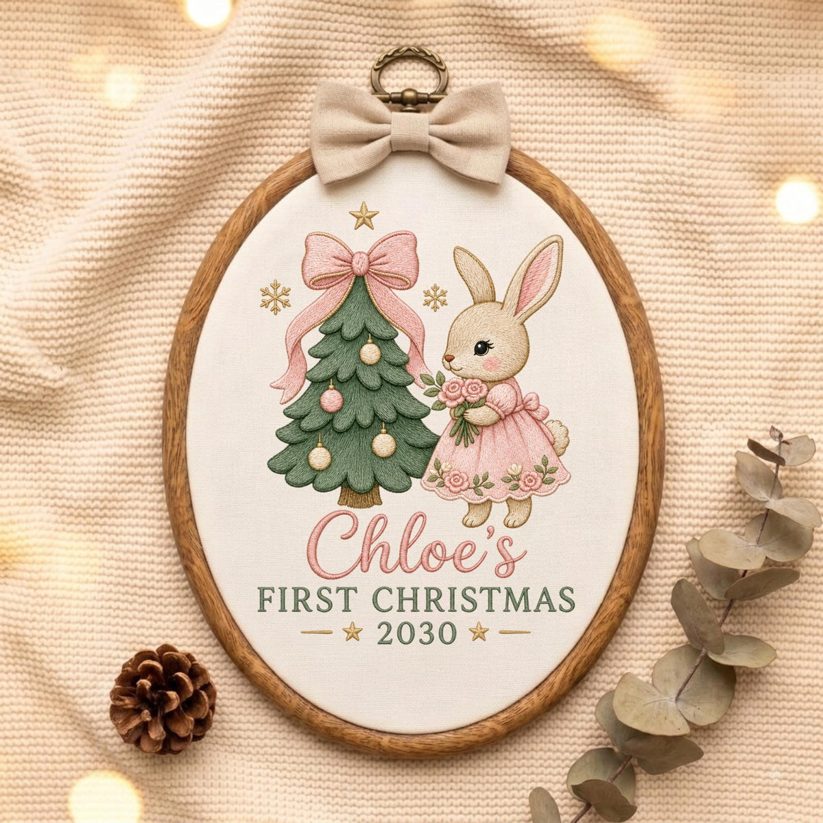 May include: An oval embroidery hoop featuring a white fabric panel with a green Christmas tree, a pink bow, and a rabbit in a pink dress. The text reads Chloe's FIRST CHRISTMAS 2030. A beige fabric bow is attached to the top of the wooden frame.