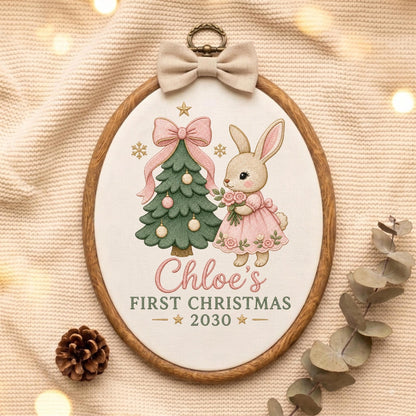 May include: An oval embroidery hoop featuring a white fabric panel with a green Christmas tree, a pink bow, and a rabbit in a pink dress. The text reads Chloe's FIRST CHRISTMAS 2030. A beige fabric bow is attached to the top of the wooden frame.