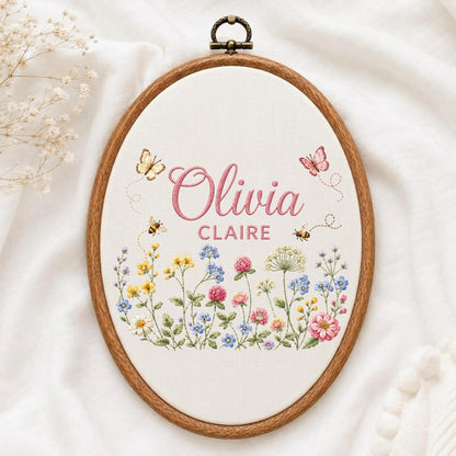 May include: An oval wooden embroidery hoop containing white fabric with the embroidered name Olivia Claire in pink script and block letters, surrounded by colorful wildflowers, small bees, and two butterflies in shades of pink and yellow.