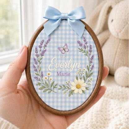 May include: An oval wooden embroidery hoop containing light blue and white gingham fabric. The fabric features embroidered lavender sprigs, white daisies, a small purple butterfly, and the names Everlyn Marie in script. A light blue satin bow is attached.