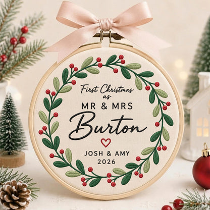 May include: A round wooden embroidery hoop containing cream fabric with a stitched green holly and red berry wreath. The text reads First Christmas as MR &amp; MRS Burton with a small red heart, JOSH &amp; AMY, and 2026. A light pink ribbon bow is attached.
