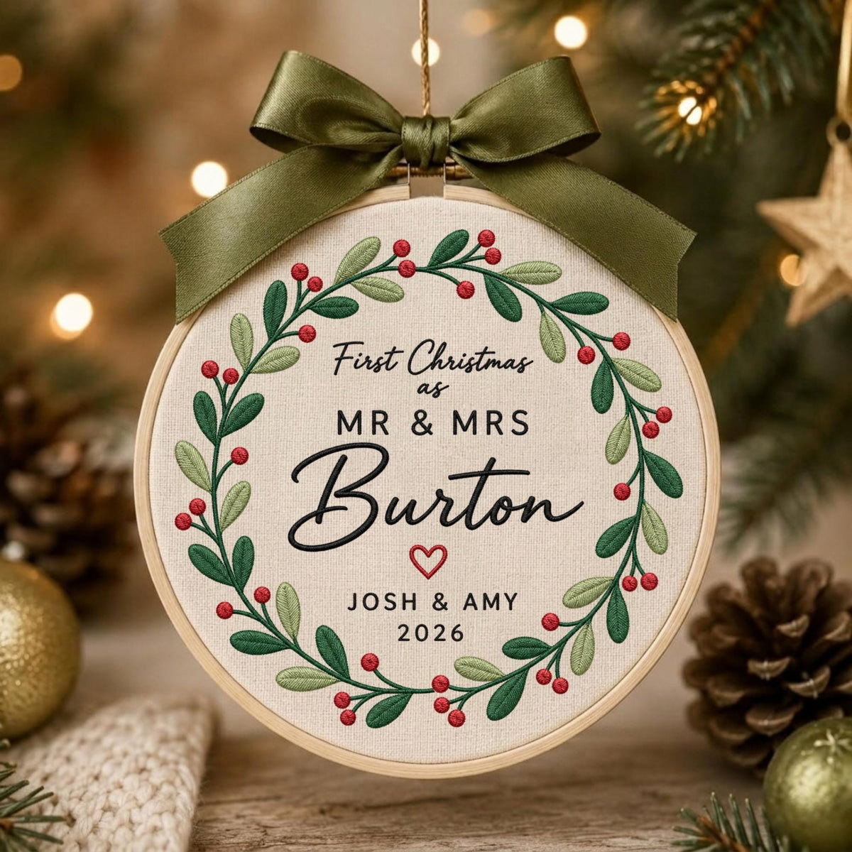 May include: A circular embroidery hoop ornament featuring a cream-colored fabric center with a green leaf and red berry wreath. The text reads First Christmas as MR &amp; MRS Burton with a small red heart, JOSH &amp; AMY, and 2026. An olive green ribbon bow.