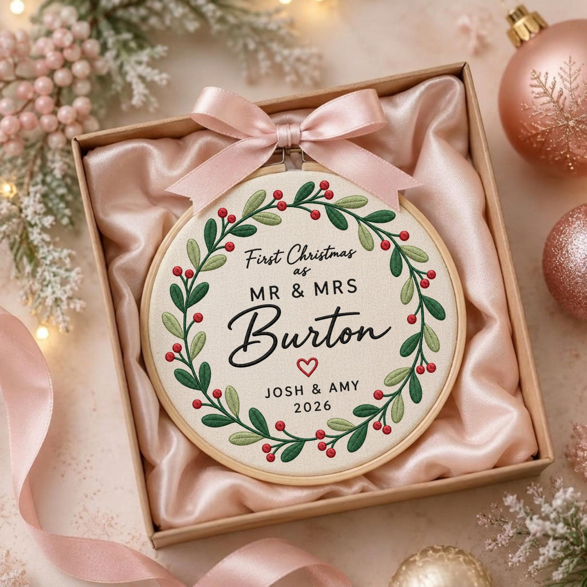 May include: A circular wooden embroidery hoop containing cream fabric with a green leaf and red berry wreath design. The text reads First Christmas as MR &amp; MRS Burton with a heart and JOSH &amp; AMY 2026. A light pink satin ribbon bow is attached.