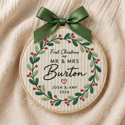 May include: A circular embroidery hoop ornament featuring a green satin bow at the top. The fabric has vertical light stripes and an embroidered wreath of green leaves and red berries. Text reads: First Christmas as MR &amp; MRS Burton, JOSH &amp; AMY, 2026.