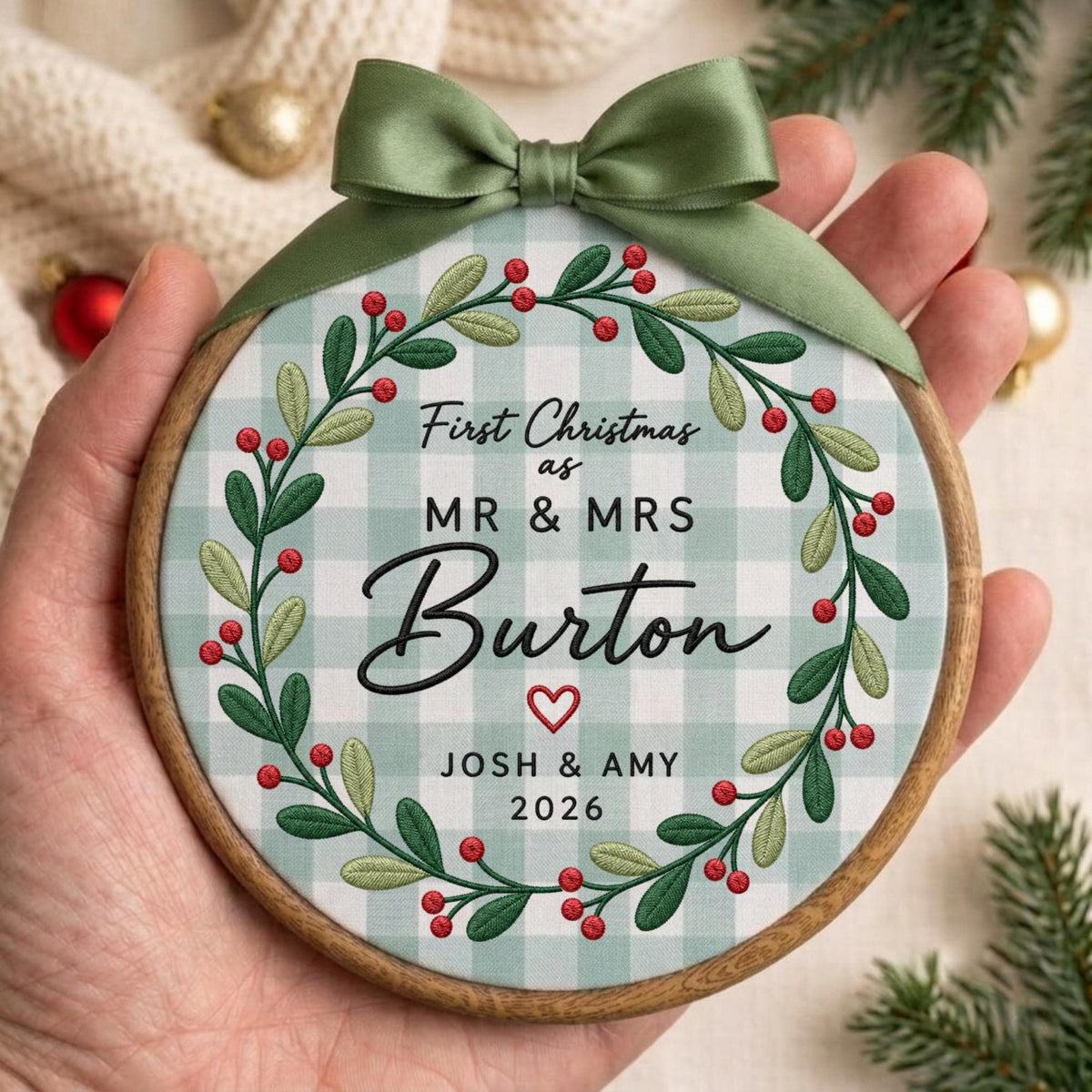 May include: A round wooden embroidery hoop ornament featuring a light green and white gingham fabric background. It displays a green leaf and red berry wreath with the text First Christmas as MR &amp; MRS Burton, a red heart, JOSH &amp; AMY, and 2026.