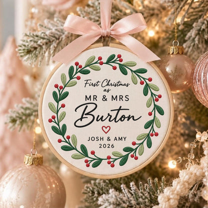 May include: A round wooden embroidery hoop ornament featuring a cream fabric center with a green leaf and red berry wreath. The text reads First Christmas as MR &amp; MRS Burton, JOSH &amp; AMY, 2026 with a small red heart. A pale pink satin ribbon bow.