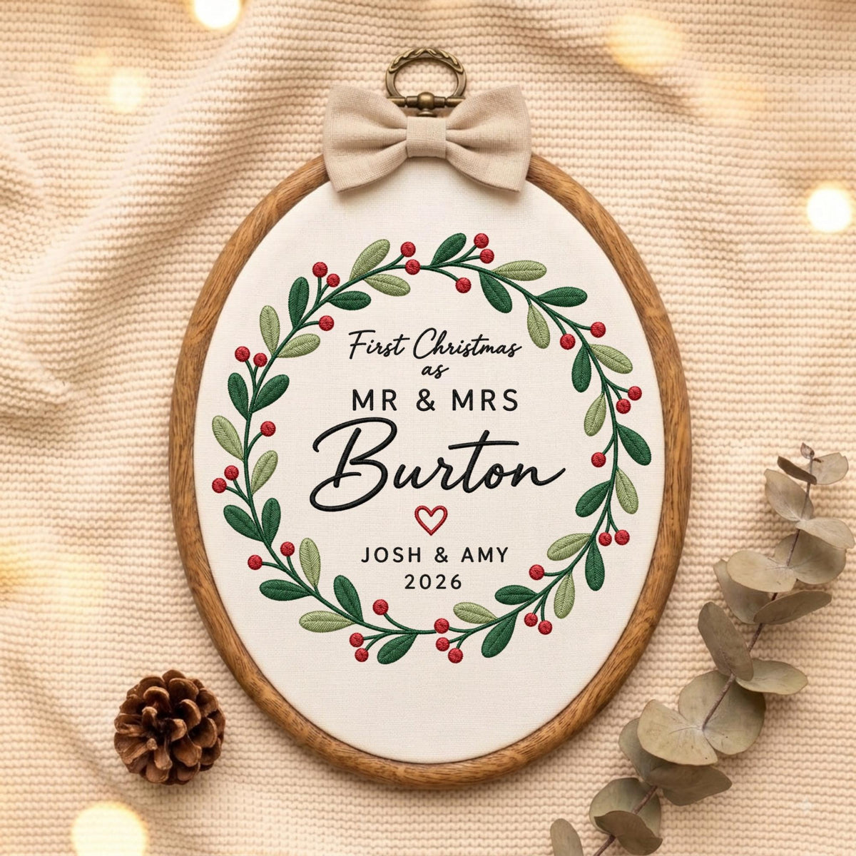 May include: An oval embroidery hoop featuring a white fabric center with a green leaf and red berry wreath design. The text reads First Christmas as MR &amp; MRS Burton with a red heart, JOSH &amp; AMY, and 2026. A beige fabric bow sits at the top.