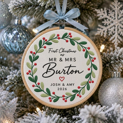 May include: A round wooden embroidery hoop ornament featuring a white fabric center with an embroidered green leaf and red berry wreath. The text reads First Christmas as MR &amp; MRS Burton with a red heart, JOSH &amp; AMY, and 2026. A light blue ribbon bow.