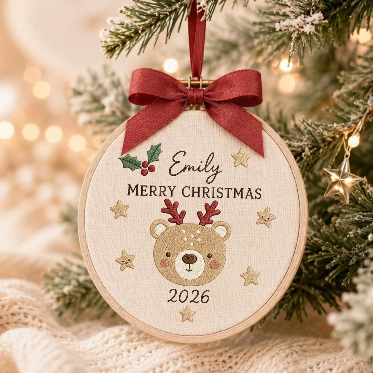 May include: A circular wooden embroidery hoop ornament featuring a beige fabric center with an embroidered brown bear face, red antlers, and the text Emily, MERRY CHRISTMAS, and 2026. It is decorated with green holly, red berries, and gold stars, topped with a dark red ribbon bow.