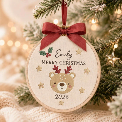 May include: A circular wooden embroidery hoop ornament featuring a beige fabric center with an embroidered brown bear face, red antlers, and the text Emily, MERRY CHRISTMAS, and 2026. It is decorated with green holly, red berries, and gold stars, topped with a dark red ribbon bow.