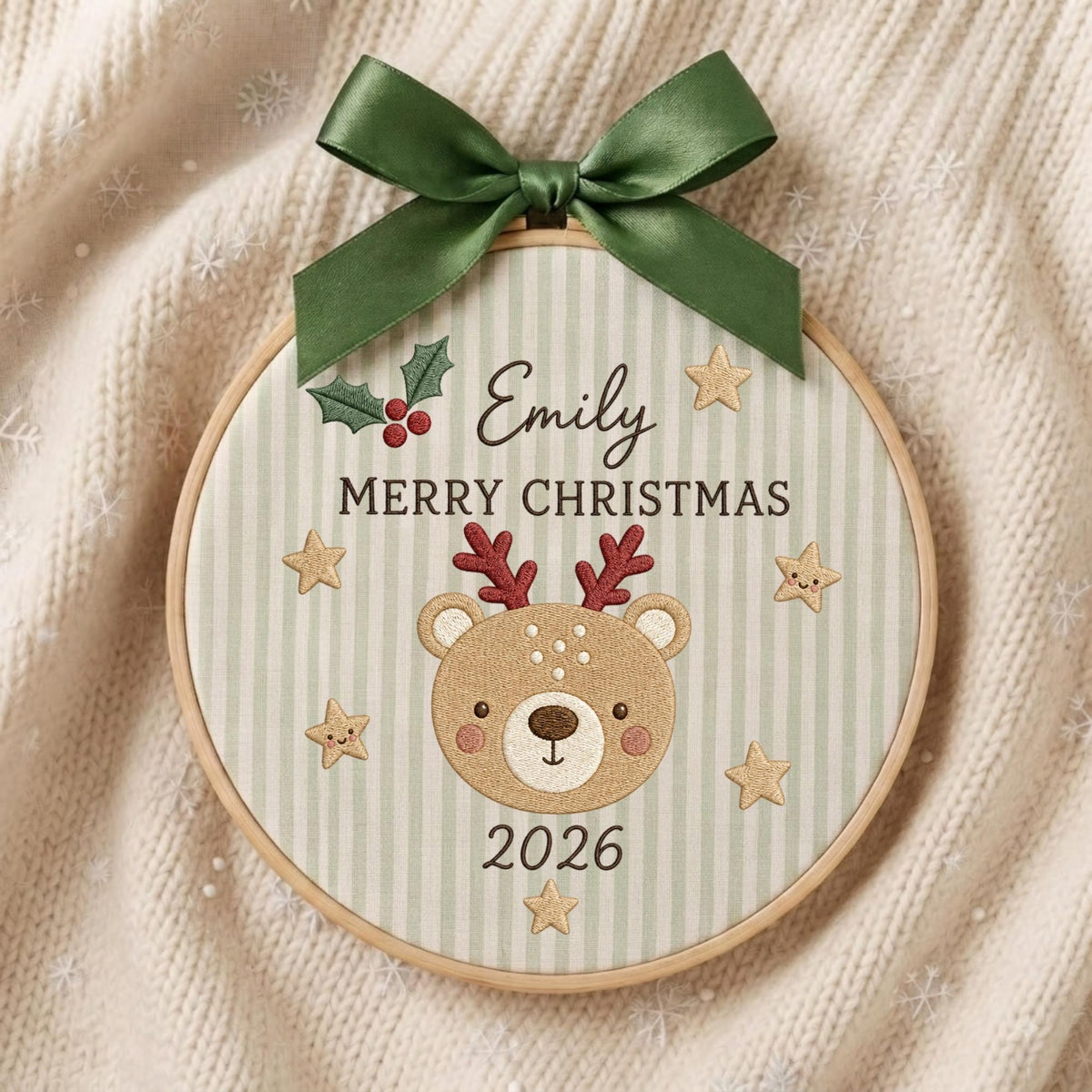 May include: A round wooden embroidery hoop ornament featuring a light green and white striped fabric background. It displays the name Emily, the text MERRY CHRISTMAS, and the year 2026. A cartoon bear with red antlers, stars, and holly berries are embroidered on the front, topped with a dark green satin ribbon bow.