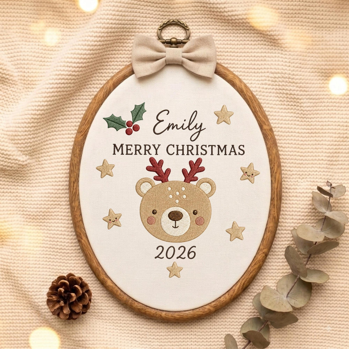 May include: An oval wooden embroidery hoop containing white fabric with an embroidered reindeer face, holly leaves, berries, and stars. The text reads Emily, MERRY CHRISTMAS, and 2026. A beige fabric bow is attached to the top metal hanging loop.