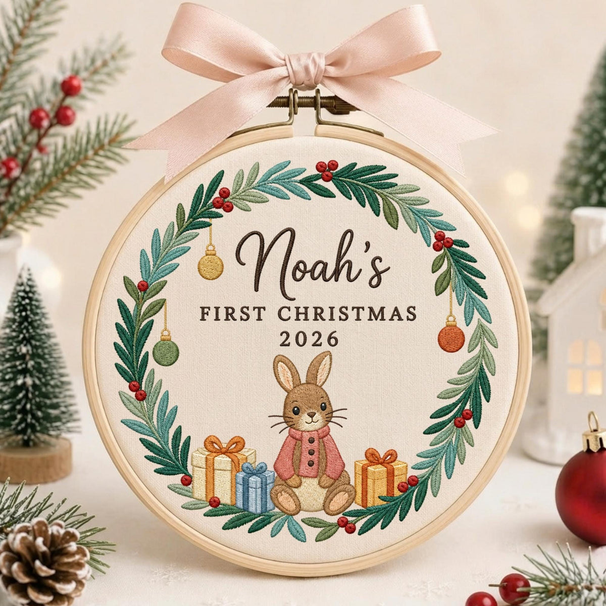 May include: A round wooden embroidery hoop containing a cream-colored fabric with a stitched wreath of green leaves and red berries. The text reads Noah's FIRST CHRISTMAS 2026 above a brown rabbit wearing a pink coat, flanked by three gift boxes.