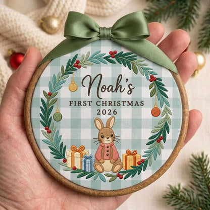 May include: A round wooden embroidery hoop ornament featuring a light blue and white gingham fabric background. A green wreath with red berries and hanging ornaments surrounds the text Noah's FIRST CHRISTMAS 2026 and a brown bunny with gifts.