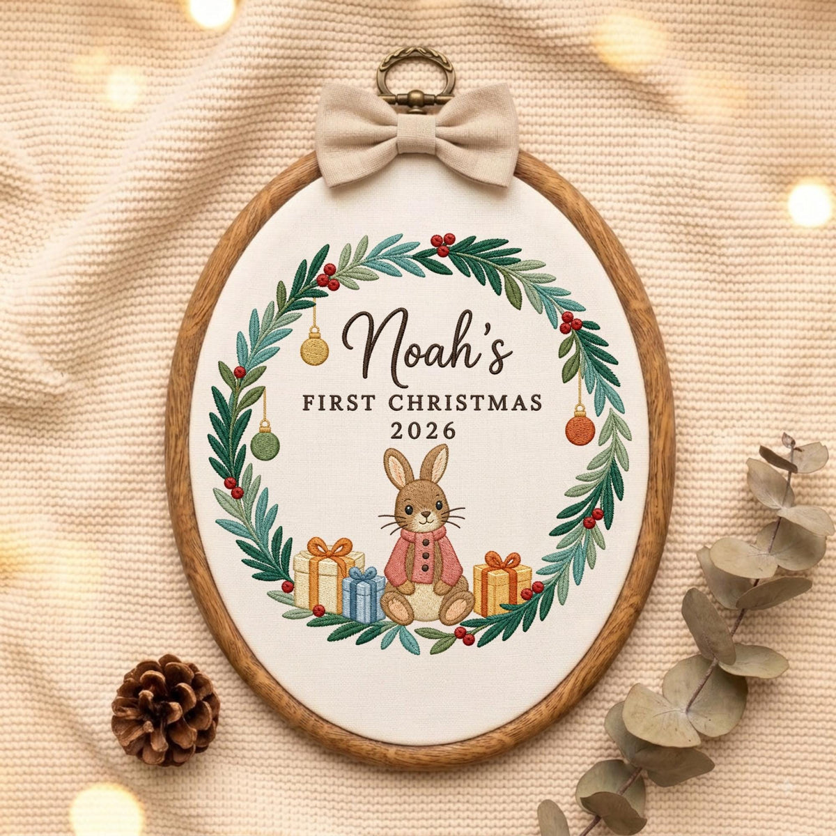 May include: An oval embroidery hoop featuring a white fabric center with a green wreath, red berries, and hanging ornaments. The text reads Noah's FIRST CHRISTMAS 2026 above a brown rabbit wearing a pink coat, flanked by two gift boxes with bows.