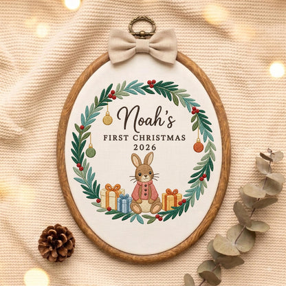 May include: An oval embroidery hoop featuring a white fabric center with a green wreath, red berries, and hanging ornaments. The text reads Noah's FIRST CHRISTMAS 2026 above a brown rabbit wearing a pink coat, flanked by two gift boxes with bows.