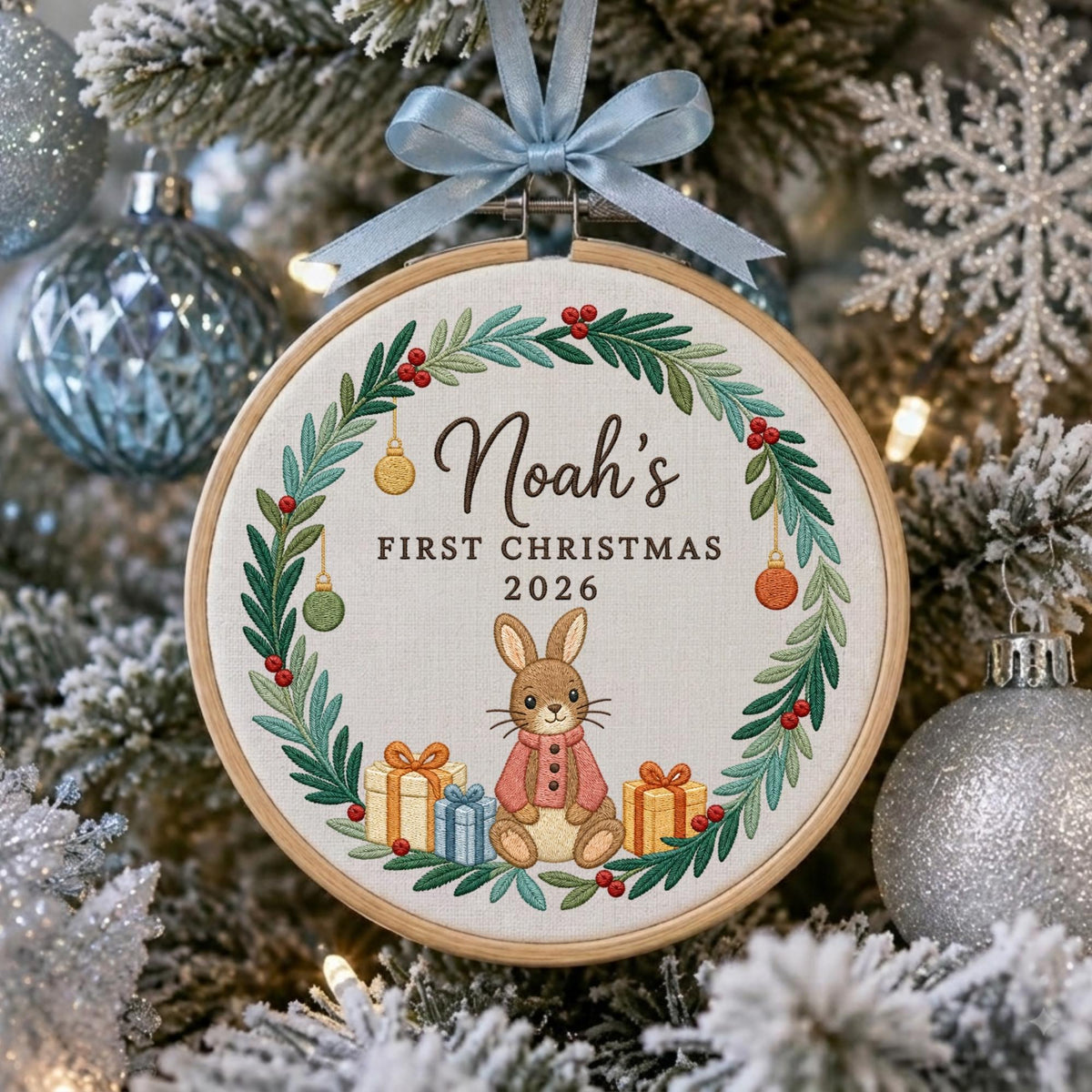 May include: A round wooden embroidery hoop ornament featuring a stitched wreath of green leaves and red berries. The text reads Noah's FIRST CHRISTMAS 2026. A brown rabbit in a pink sweater sits among gift boxes, topped with a light blue ribbon bow.