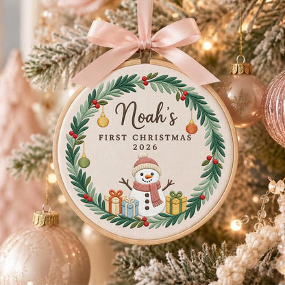 May include: A round embroidery hoop ornament featuring a green pine wreath with red berries and hanging ornaments. In the center, a snowman wears a pink hat and scarf, surrounded by gift boxes. Text reads: Noah's FIRST CHRISTMAS 2026.