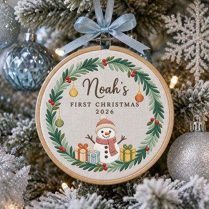 May include: A circular embroidery hoop ornament featuring a green pine wreath with red berries and hanging ornaments. In the center, a snowman wears a pink scarf, flanked by two gift boxes. Text reads: Noah's FIRST CHRISTMAS 2026. A light blue ribbon bow is attached to the top.