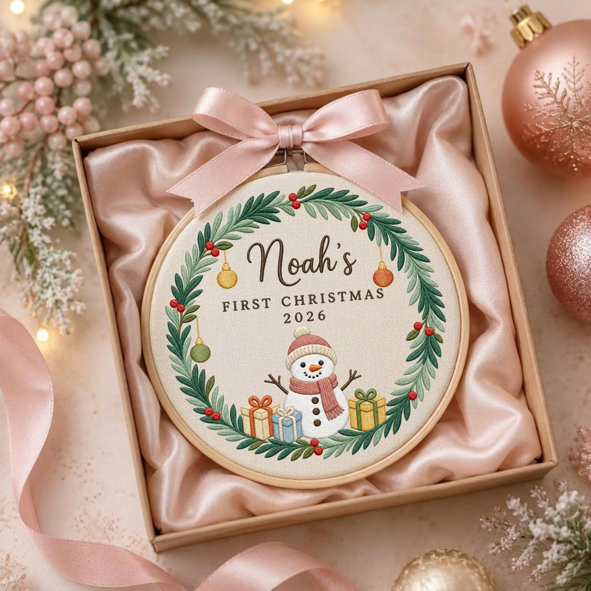 May include: A round wooden embroidery hoop containing fabric with a stitched wreath of green leaves and red berries. The center features the text "Noah's FIRST CHRISTMAS 2026" above a snowman with gifts. A light pink satin ribbon is tied at the top.