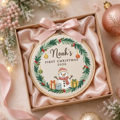 May include: A round wooden embroidery hoop containing fabric with a stitched wreath of green leaves and red berries. The center features the text "Noah's FIRST CHRISTMAS 2026" above a snowman with gifts. A light pink satin ribbon is tied at the top.